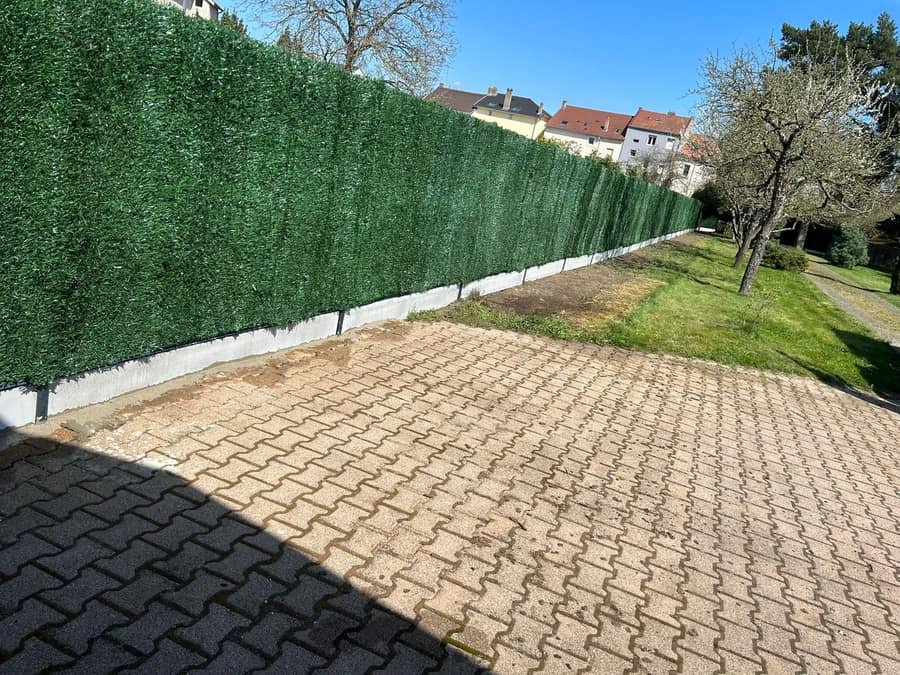 Haie artificielle brise-vue installee sur un grillage existant le long d'une cour pavee